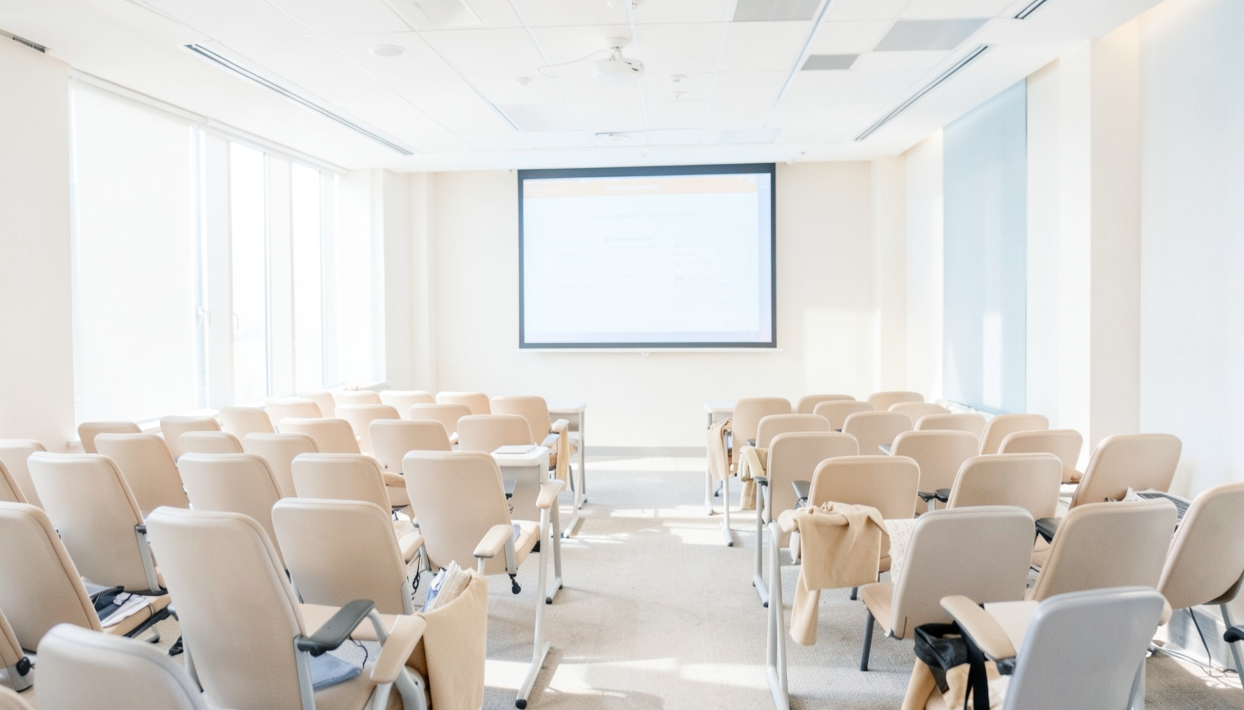 A bright, airy conference room with rows of seating and a presentation screen, bathed in natural daylight with neutral beige and soft blue tones.